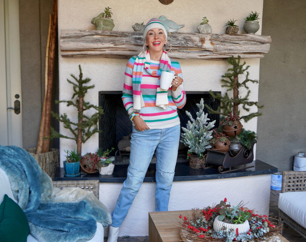 Sheree Frede of the SheShe Show sitting at outdoor fireplace wearing a hot pink vest and colorful sweater, scarf and hat