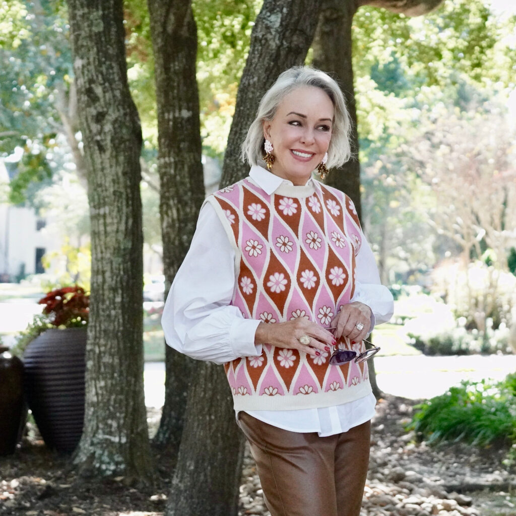 Sheree Frede of the SheShe Show standing in the trees wearing a pink sweater vest with white shirt over faux leather pants