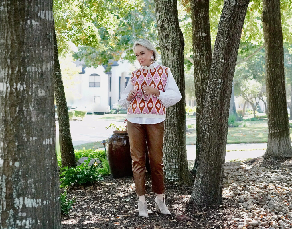 Sheree Frede of the SheShe Show standing in the trees wearing a pink sweater vest with white shirt over faux leather pants