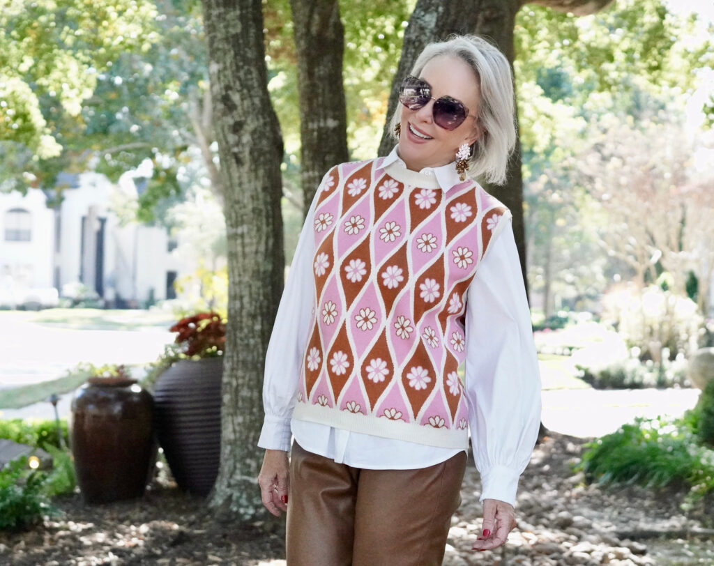 Sheree Frede of the SheShe Show standing in the trees wearing a pink sweater vest with white shirt over faux leather pants