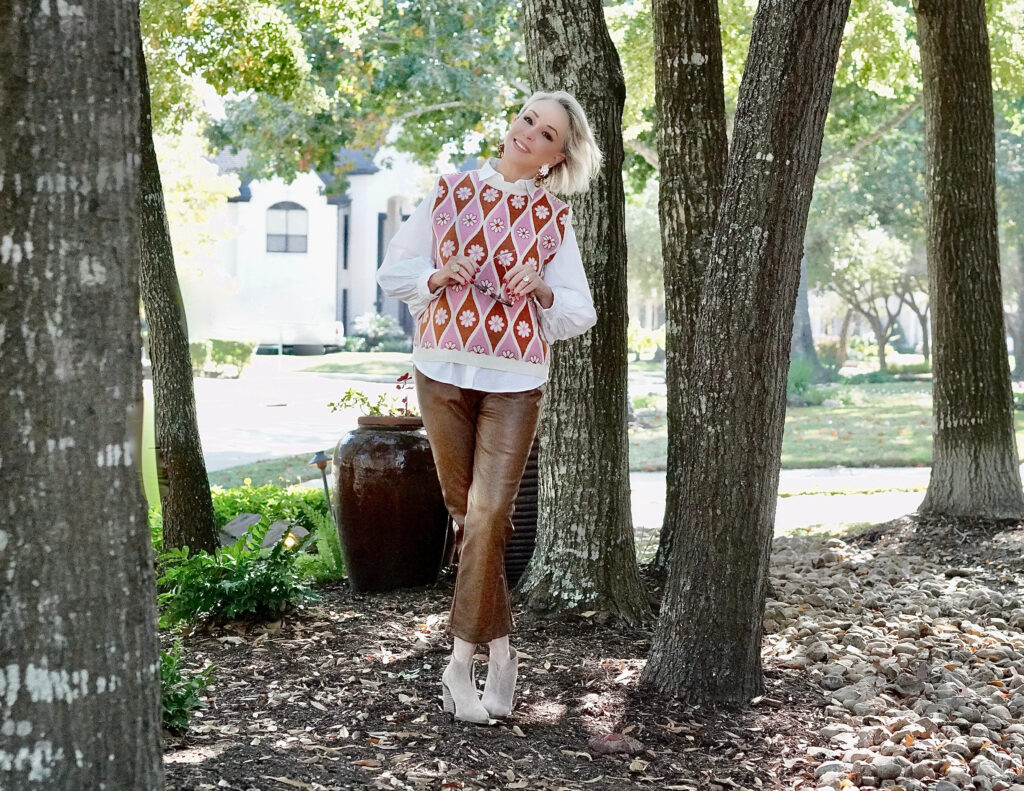 Sheree Frede of the SheShe Show standing in the trees wearing a pink sweater vest with white shirt over faux leather pants