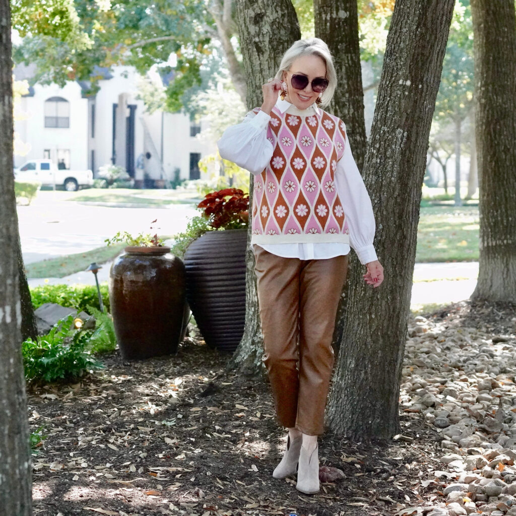 Sheree Frede of the SheShe Show standing in the trees wearing a pink sweater vest with white shirt over faux leather pants