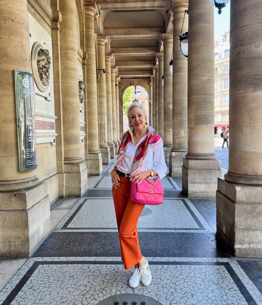 Sheree standing in arched columns wearing orange faux leather crop pants and Chico's no-iron white button front shirt holding a hot pink Chanel handbag