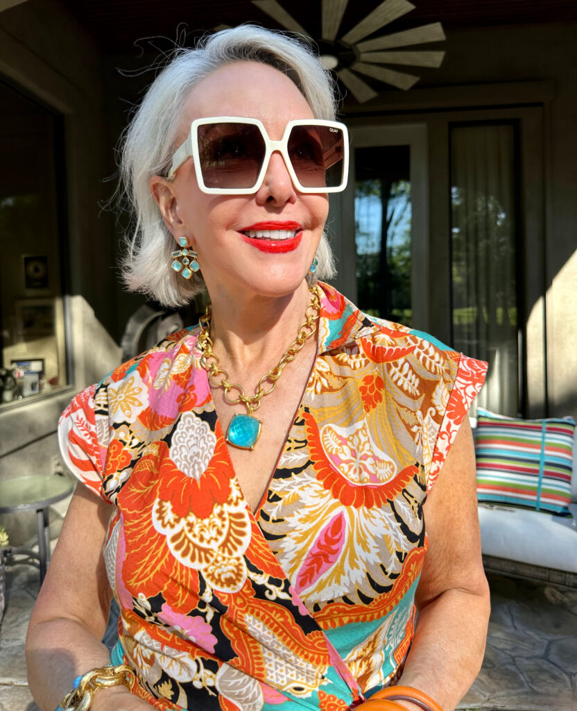 Sheree Frede wearing an orange print dress and big white sunglasses with turquoise and gold pendant and earrings by Julie Voss
