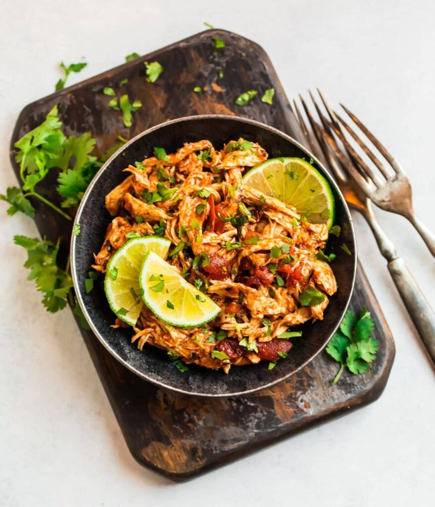 This photo is a Cinco de Mayo meal including bowl of slow cooker Mexican chicken garnished with fresh limes and cilantro sitting atop a wooden cutting board and forks.