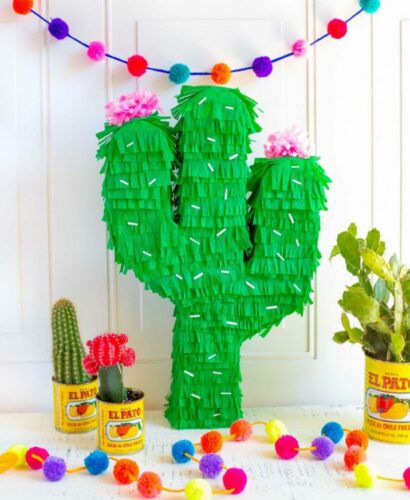This is a photo of a cactus pinata, colorful pom pom garland and 3 mini cacti potted in metal canisters.