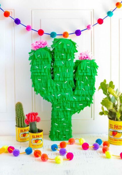 This is a photo of a cactus pinata, colorful pom pom garland and 3 mini cacti potted in metal canisters.