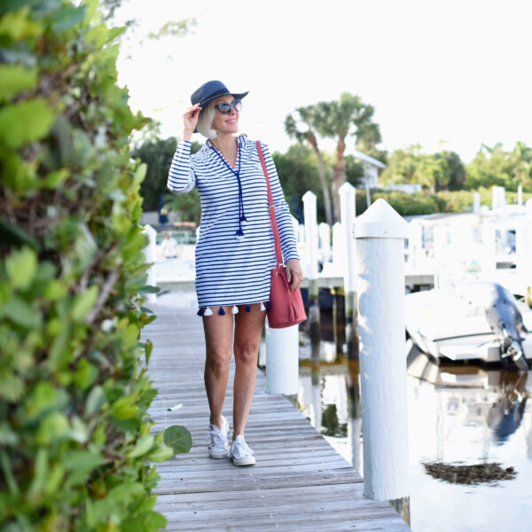 Sheree Frede of the SheShe Show on a pier dock wearing a blue and white Cabana Life sundress, red purse, white sneakers and navy hat with sunglasses displaying a casual Memorial Day look.