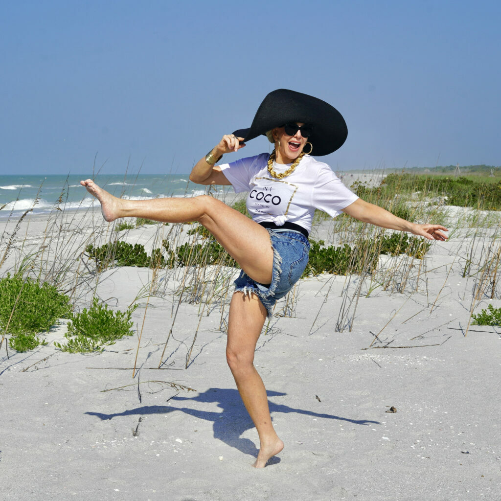 Photo of Sherre Frede (SheShe) on the beach in a Coco Chanel shirt with her leg kicked in the air and a black floppy beach hat showing how to wear shorts over 40 years old.