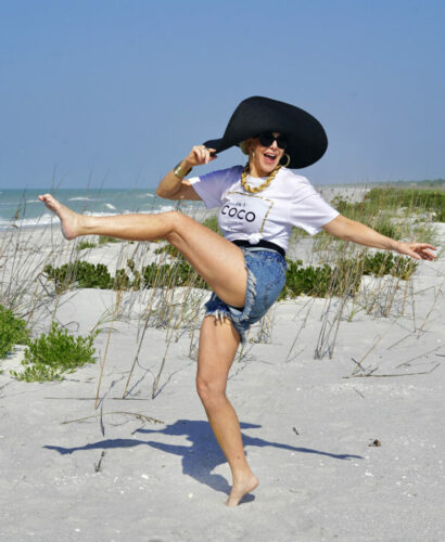 Photo of Sherre Frede (SheShe) on the beach in a Coco Chanel shirt with her leg kicked in the air and a black floppy beach hat showing how to wear shorts over 40 years old.