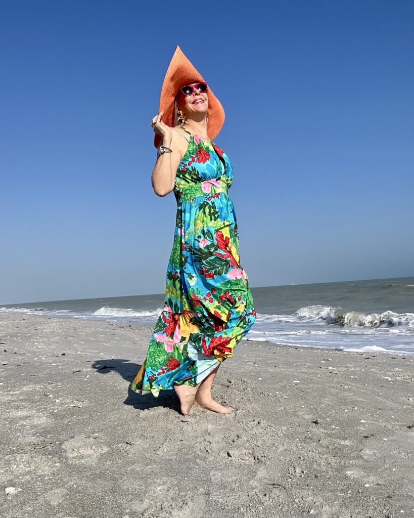 Sheree Frede of SheSheShow in floral dress and orange floppy hat on beach for resort wear