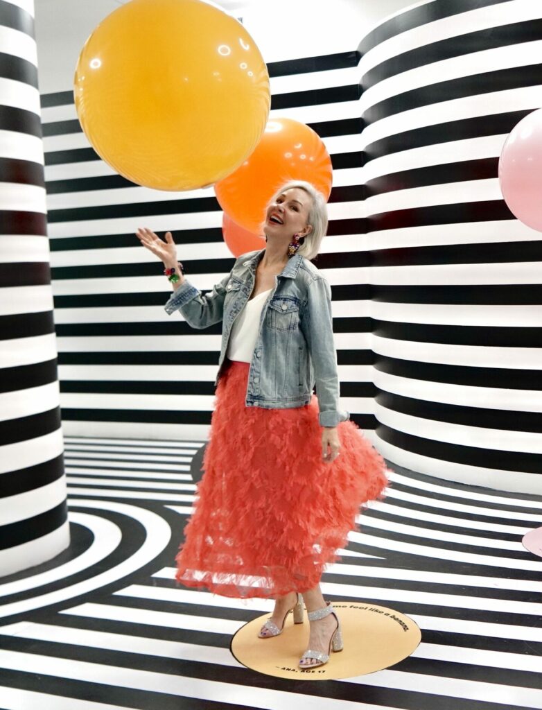 Sheree Frede wearing a coral tulle skirt with denim jacket in a black and white stripe room holding a yellow and orange balloon